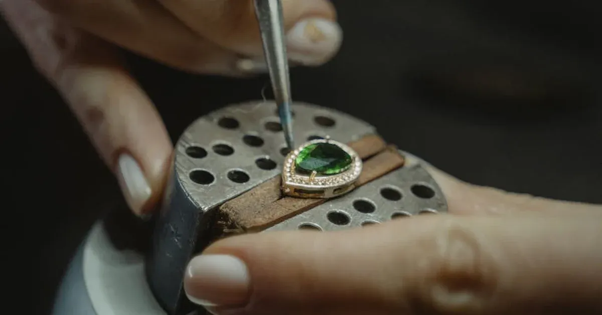 Close-up of a jeweler&#x27;s hands meticulously setting a vibrant green gemstone into an ornate gold ring, surrounded by tiny diamonds, on a specialized workbench.