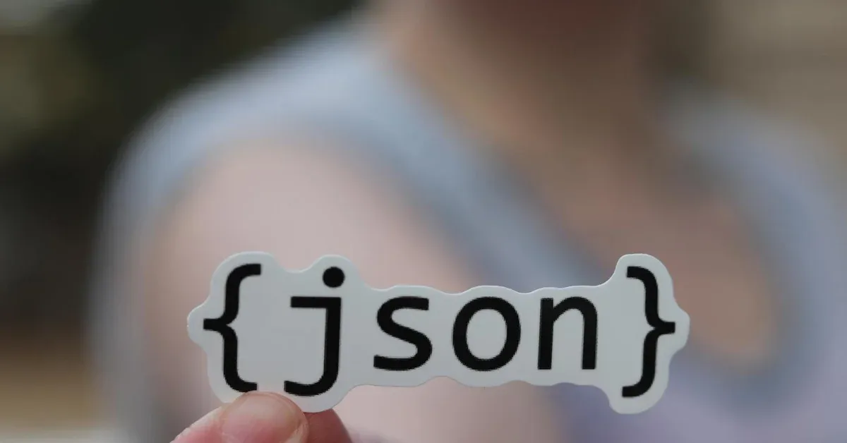 A hand holds a white sticker with the black text "{json}", symbolizing a new, faster digital data format for comics.