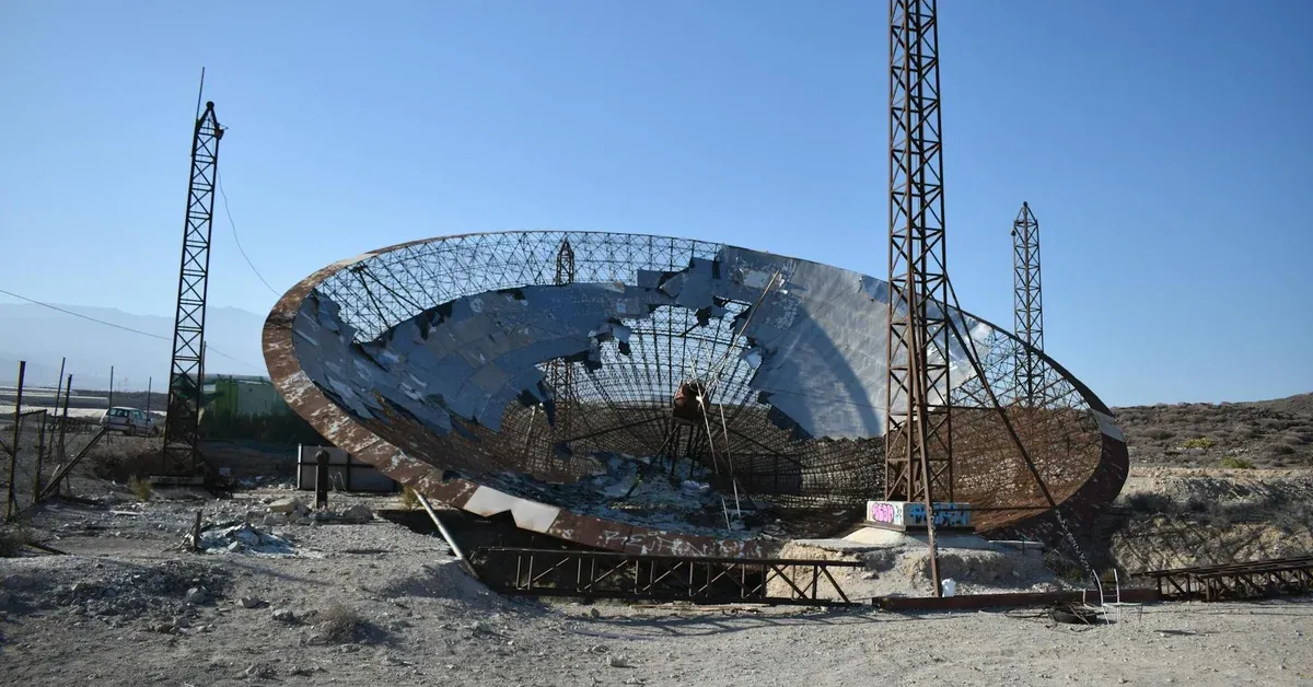 A massive, partially collapsed and rusted satellite dish lies abandoned in a stark, arid landscape under a clear sky, symbolizing decaying digital infrastructure.