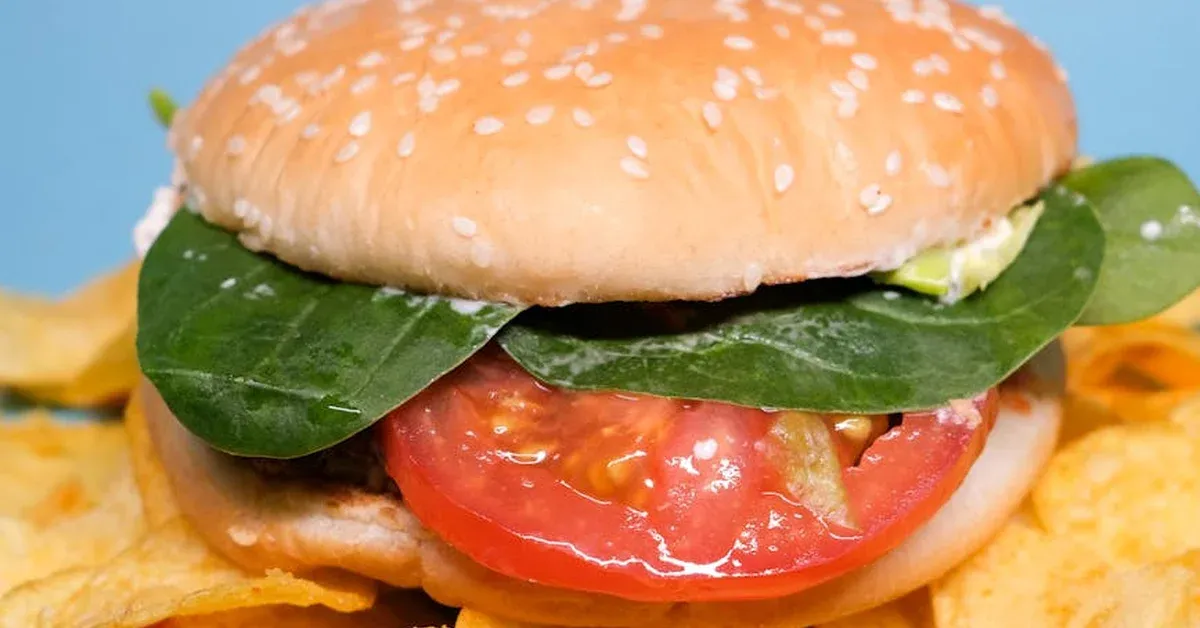 A close-up of a sesame seed bun hamburger with fresh spinach and a thick tomato slice, symbolizing the link between food and the fatal Alpha-Gal meat allergy.