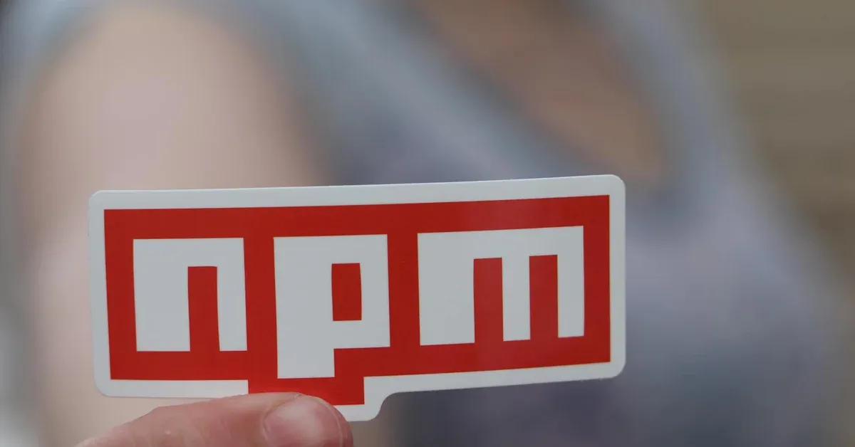 A hand holds an npm (Node Package Manager) logo sticker, a widely recognized symbol of the open-source JavaScript ecosystem.